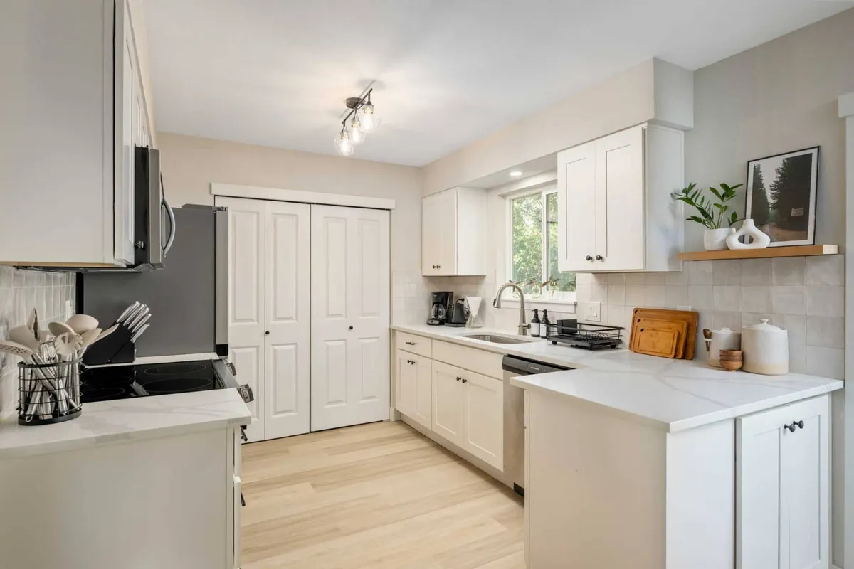 This image showcases a bright, modern kitchen with a prominent kitchen island, contemporary appliances, and shelves adorned with tasteful decor. Natural light streams in through a large window, emphasizing the welcoming and open nature of the space. The kitchen appears ready for cooking, socializing, and family gatherings.