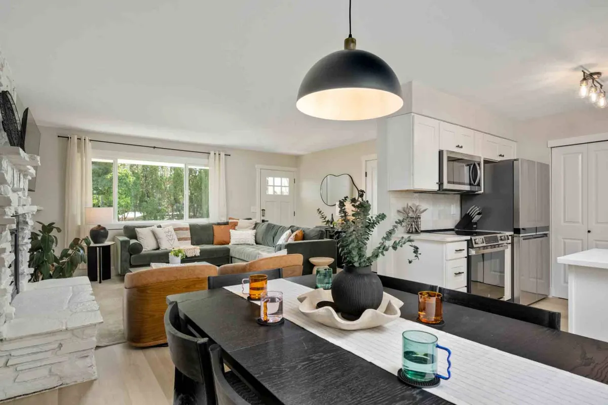 This image features a spacious living and dining area bathed in natural light, with a large sectional couch, a welcoming stone fireplace as the centerpiece, and a stylish dining table set with colorful glasses. The open floor plan allows for seamless movement between lounging and dining, while the large windows invite leafy outdoor views into the space.