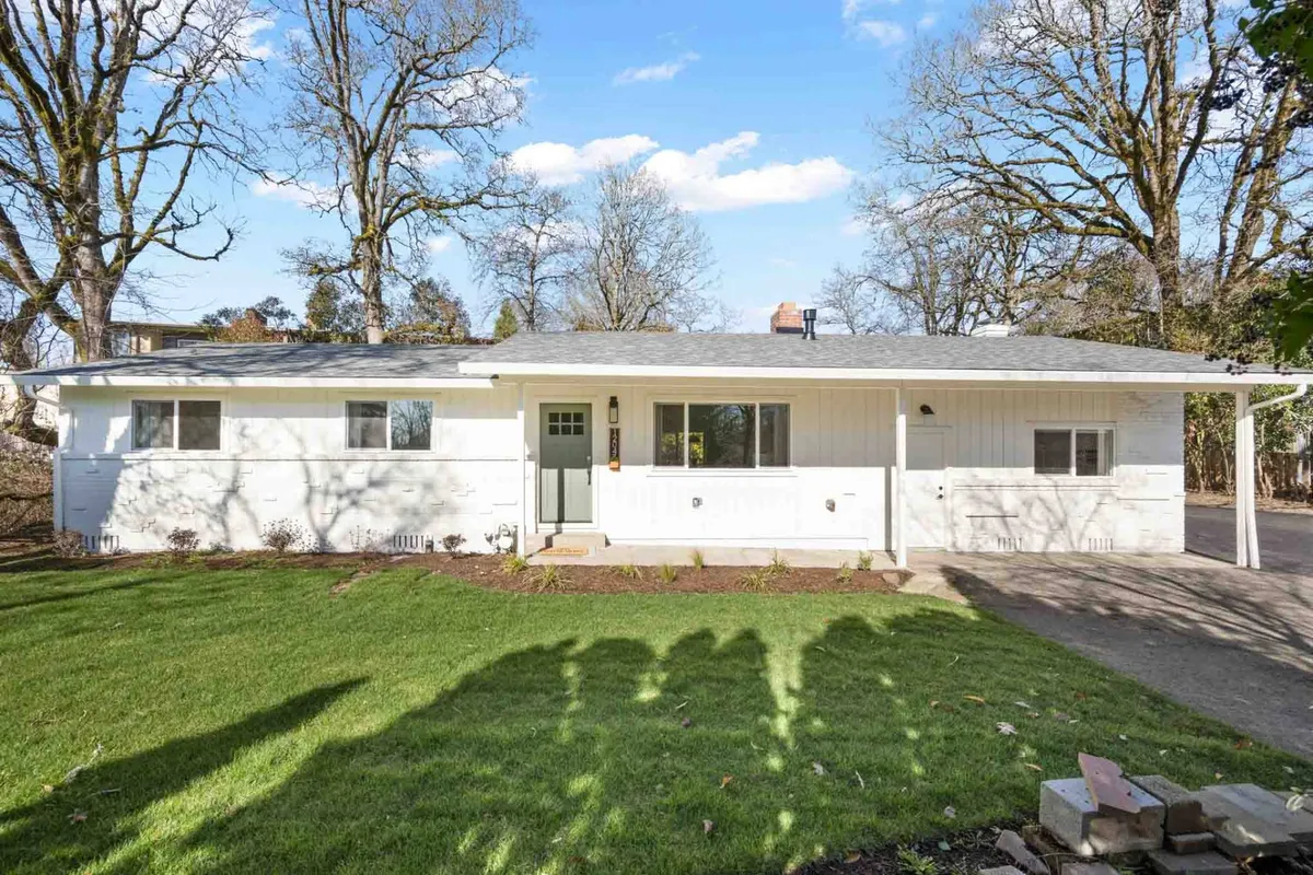 The image captures the inviting white exterior of The Holistic Hideaway, with its well-maintained lawn and mature surrounding trees. The front door is visible, and the setting is tranquil, suggesting a peaceful retreat close to nature.