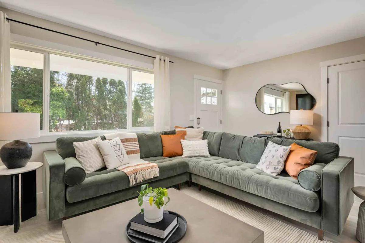 A green sectional sofa is oriented to enjoy the natural light streaming through a large window, which frames tranquil views of greenery outside. Decorative pillows and a modern coffee table create a cozy, inviting spot for reading, relaxing, or socializing gently with friends or family.