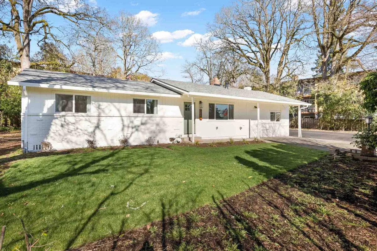 The modern white facade of the home overlooks a vibrant, well-kept yard. Large windows and a welcoming porch area further connect the living space to the outdoors, while the expansive grass is perfect for pets or children to play. The landscaping is neat and thoughtfully designed, reinforcing the sense of privacy and calm.