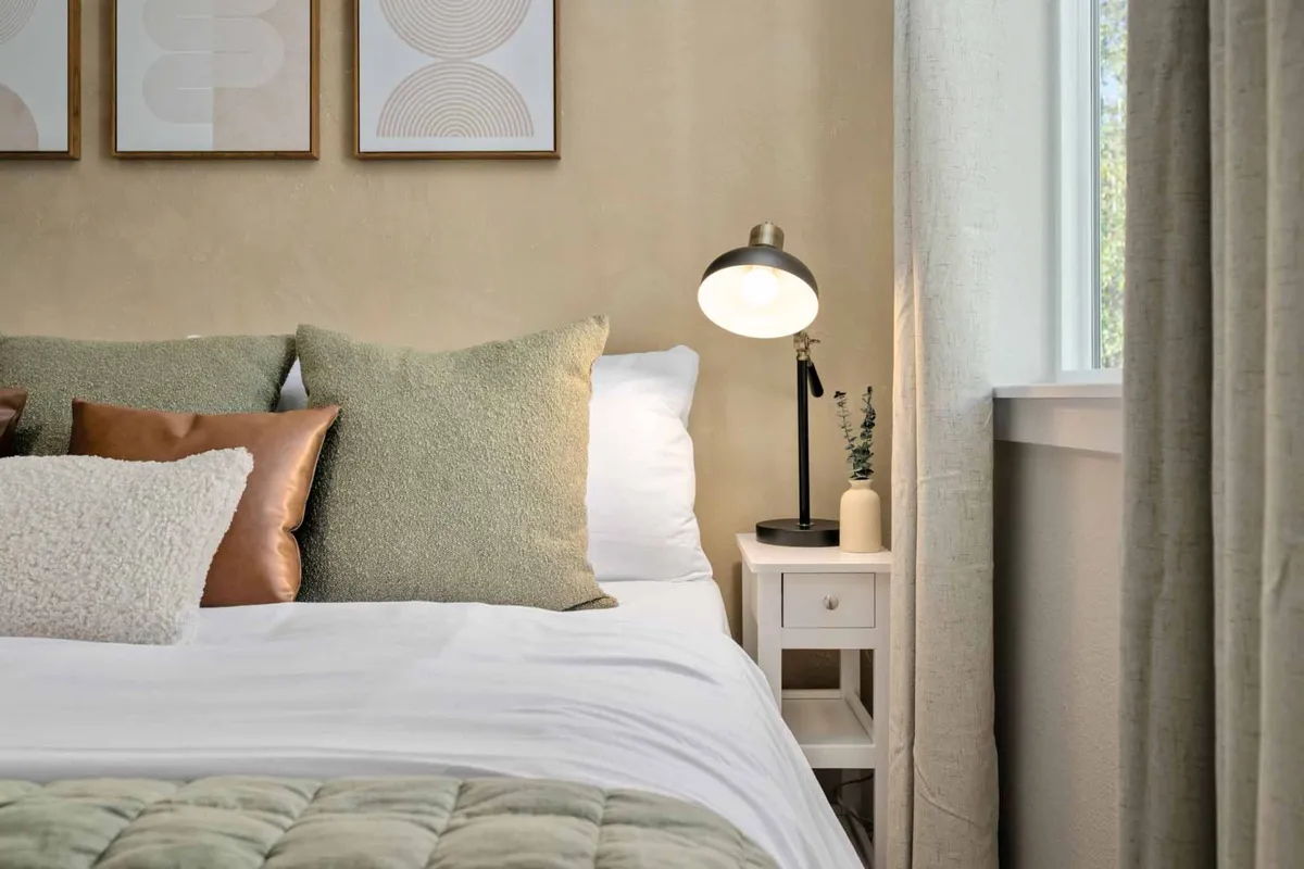 A cozy bedroom with a stylish bed adorned with decorative pillows, a nightstand featuring a lamp, and artwork on the wall. The gentle lighting and inviting color palette contribute to a restful, comfortable space.