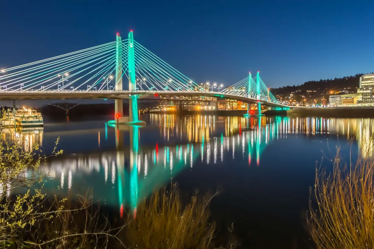 A vibrant evening cityscape shows a suspension bridge, water reflections, and glowing city lights, hinting at bustling nightlife and scenic dining possibilities along the waterfront.