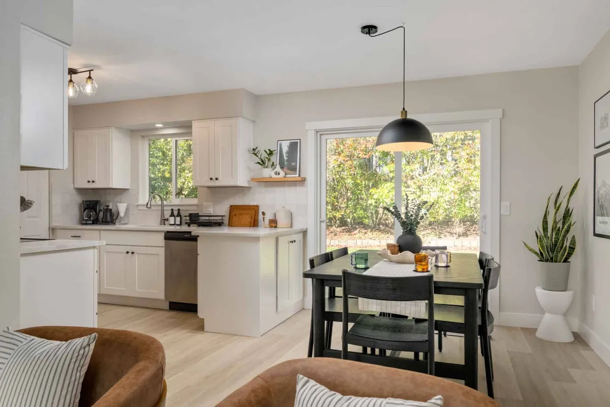 A warm and welcoming dining area with a large table set near a window offering a view to the outside. The space flows seamlessly into a kitchen area and comfortable seating space, suggesting communal meals and morning routines. Natural light streams in, creating an inviting atmosphere ideal for winding down after a busy day.
