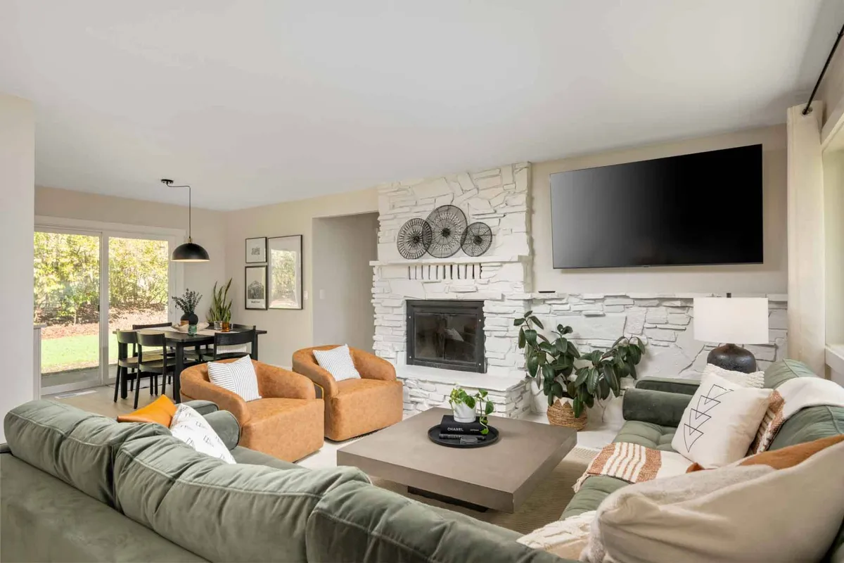 A warm, inviting interior shot shows a plush sofa facing a prominent fireplace, with a dining table positioned nearby. Decorative wall art and a wall-mounted TV add homely touches, while the room's open layout fosters social interaction and relaxation. Ample light enters through sizeable windows, creating a comfortable, family-friendly environment.