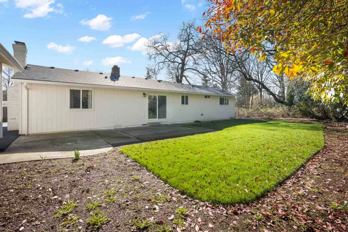 A tranquil, well-maintained lawn stretches out behind the house. Clean white siding and large windows create a bright, inviting look, while mature landscaping and trees surround the yard, offering privacy and a natural retreat. The patio area is ideal for morning coffee or relaxing with pets.