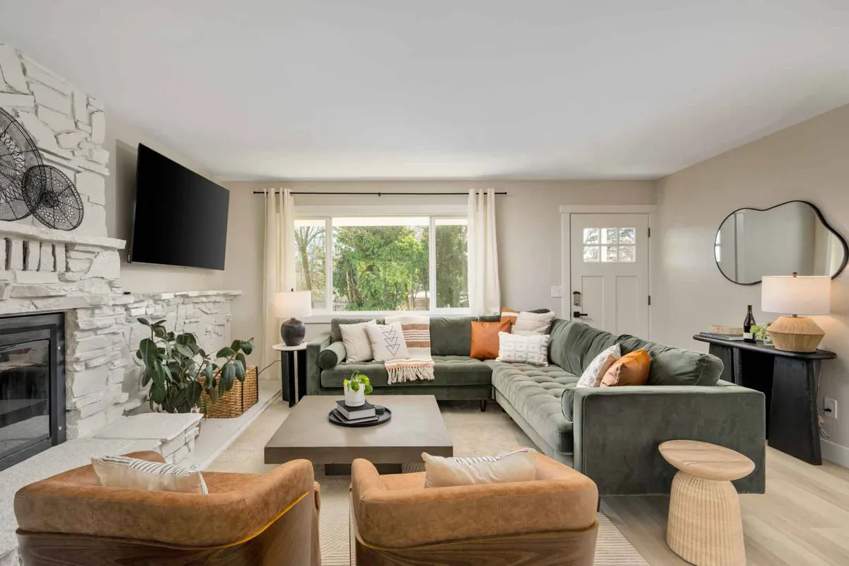 The image showcases a spacious, light-filled living room featuring a large sectional sofa centered around a crackling fireplace, a modern coffee table, and lush decorative plants. Expansive windows frame the greenery outside, blending indoor comfort with natural beauty. The space radiates warmth and relaxation, inviting guests to gather, unwind, or enjoy quiet moments.
